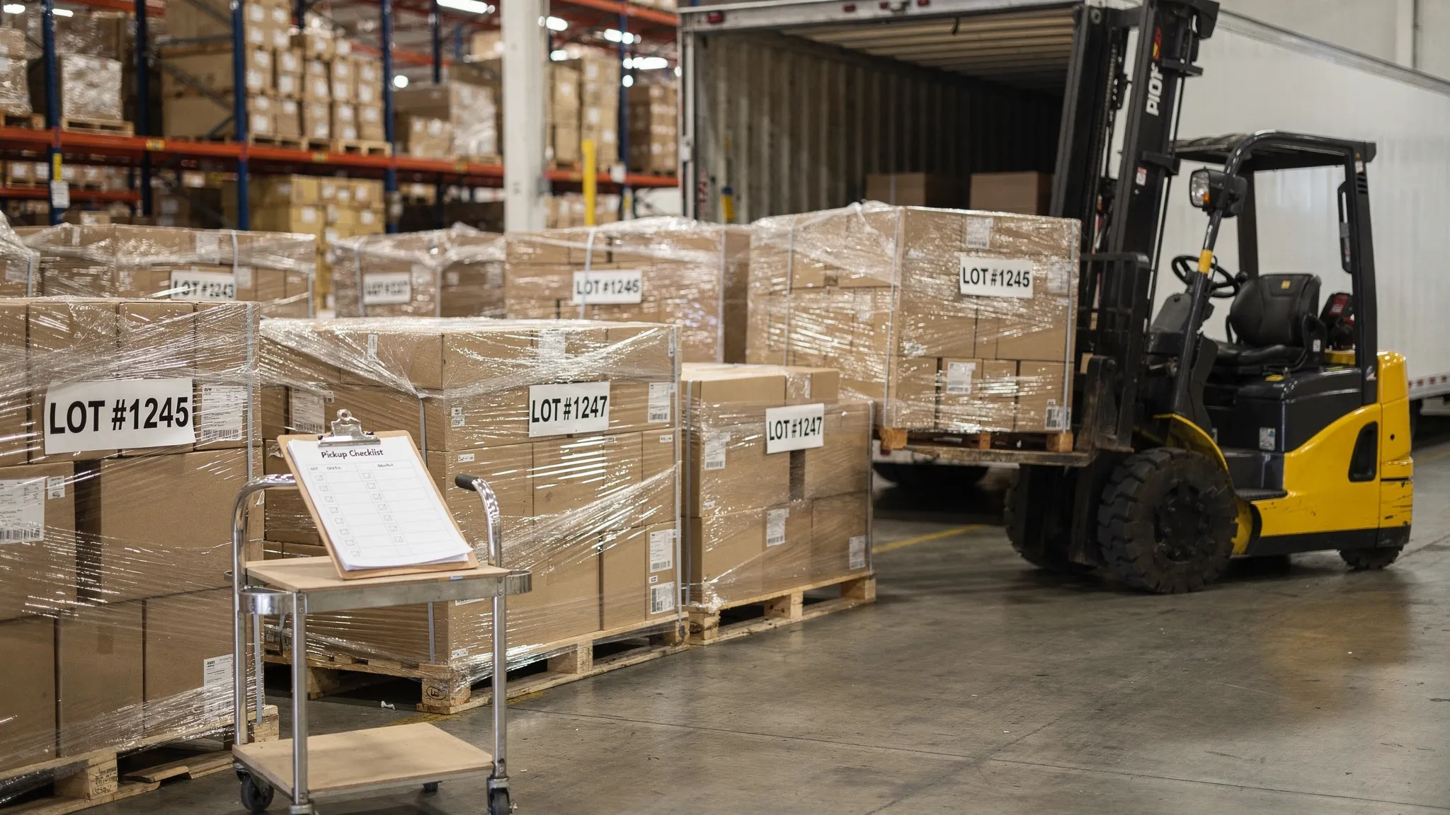 A reseller warehouse staging area with several shrink-wrapped liquidation pallets labeled by lot number, a forklift positioned to load a pallet into an open trailer, and a clipboard showing a simple pickup checklist.