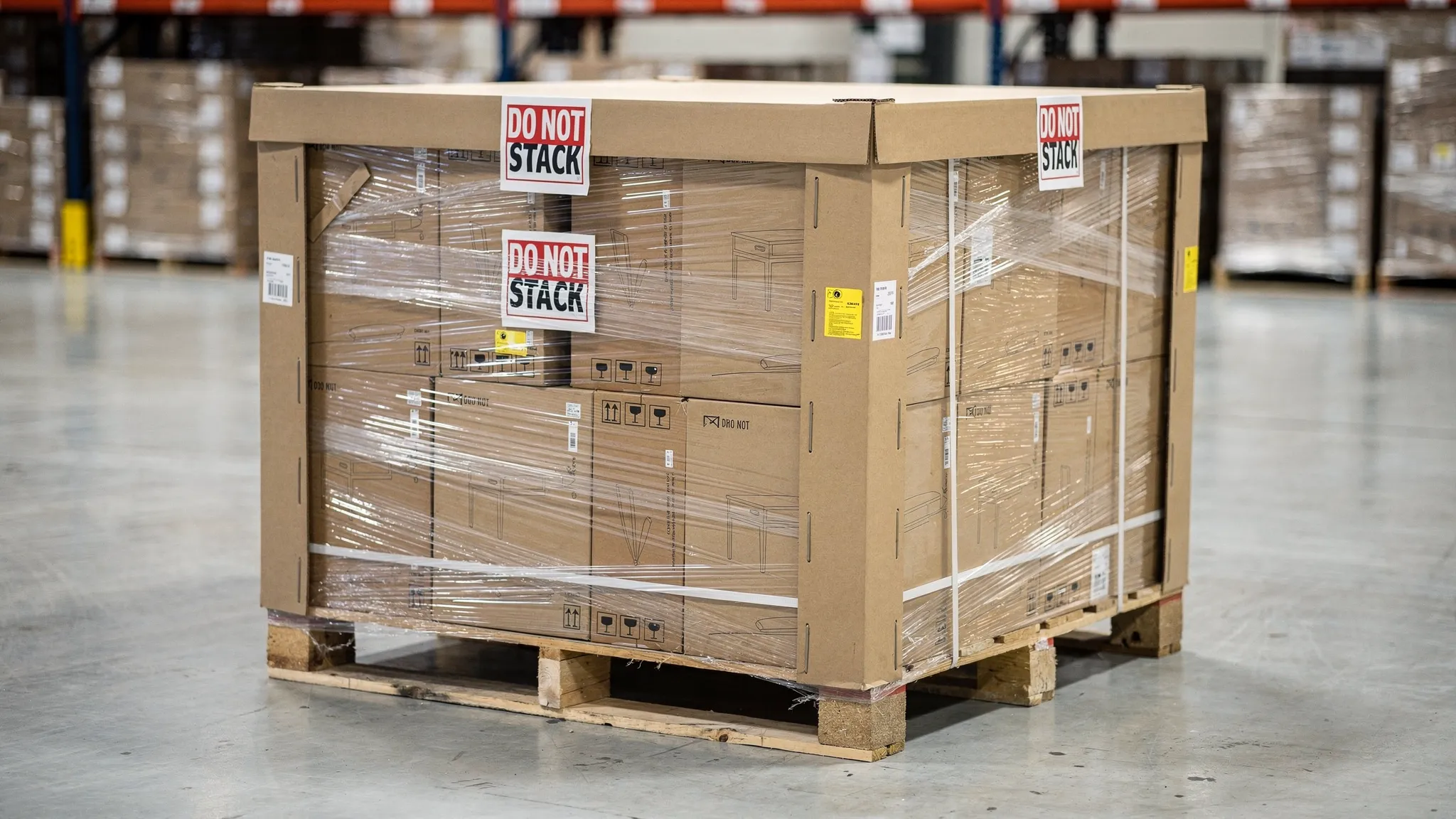 A wrapped and banded pallet of boxed furniture on a warehouse floor, showing corner protectors, a top cap, “Do Not Stack” labels, and clearly visible pallet edges with no box overhang.
